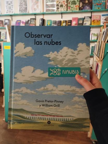 Portada del libro "Observar las nubes": un paisaje con cielo azul y nubes blancas. Las letras que componen el título y sus autores están en color negro. Aparece también uno de nuestros marcapáginas de Naubis