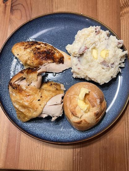 Photo of a blue plate loaded with slices of chicken, red mashed potatoes, and a dinner roll with butter on top of it.