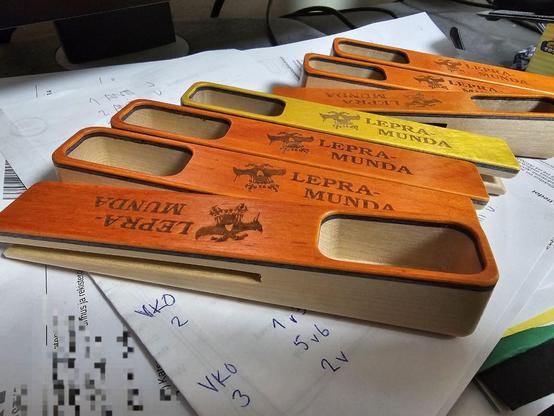 Kahvimittoja sekalaisen paperikasan päällä. Mitat on valmistettu koivusta, jonka päällä ohut musta viilu, jonka päällä ohut oranssi viilu. Oranssiin kädensijaan laseroitu teksti "LEPRAMUNDA" sekä tyylitelty kallon kuva. Yhdessä mitassa oranssin tilalla keltainen viilu.