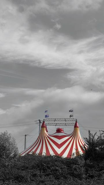 Ein leuchtend rot-weiß gestreiftes Zirkuszelt steht vor einem monochromen Himmel, mit sichtbaren Fahnen auf der Oberseite der Struktur. Das umliegende Grün ist in der Farbe gedämpft, was das Zelt betont.