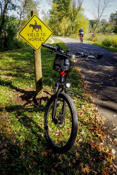 A black and red bicycle in front of a sign that says "Yield to horses" along a trail through the woods. Another biker is visible on the trail in the background.