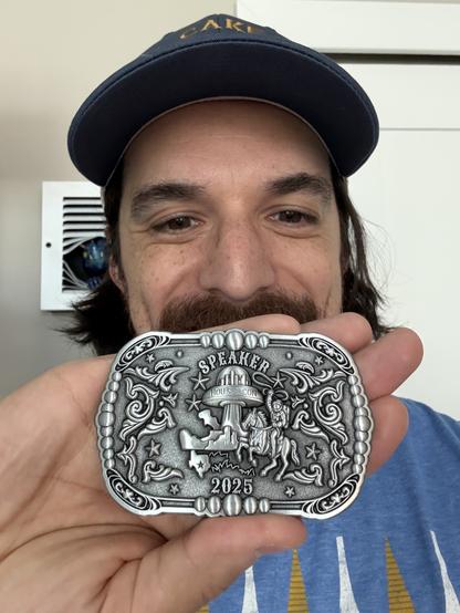 Selfie of Jimmy holding a belt buckle. The belt buckle is a western style buckle. The buckle has Speaker along the top, an image of the HOUSECCON flying saucer logo below it, and an astronaut riding a horse. The bottom of the buckle has the year, 2025. The rest of the buckle is decorated with filigree.
