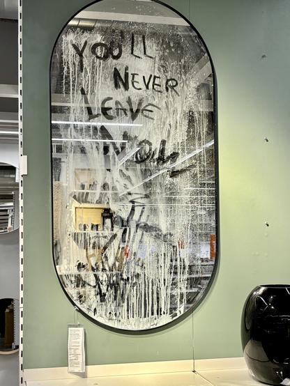 Photo of a mirror in an IKEA store. The mirror is very dirty, with the grey dirt in rivulets down the mirror. Someone has marked into the dirt with their finger: "you'll never leave now"