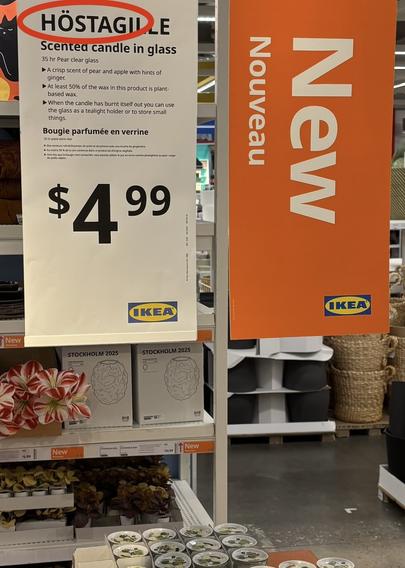 Photo of a large hanging product sign, in white, with another hanging sign in orange explaining "New". The white sign says "Höstagile, scented candle in glass, $4.99" but I've added a red oval around the part that says "Höstag", because there is clearly a subliminal message.
