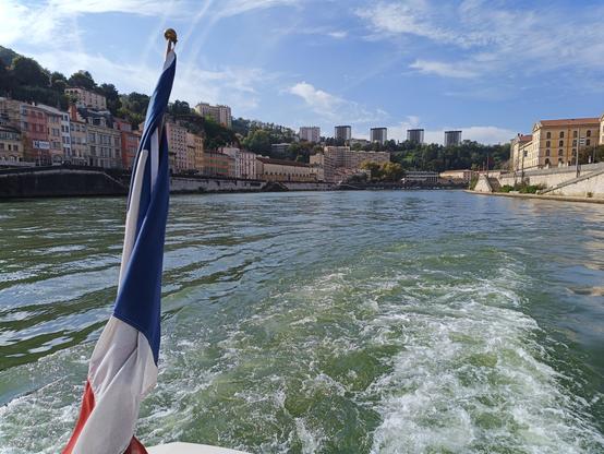 4 photos numériques couleur en format paysage, avec une vue prise à l'arrière d'un bateau qui navigue sur une rivière large. Il y a un drapeau français enroulé par le vent, les remous sur l'eau, des immeubles de part et d'autre avec à gauche une colline boisée. Le ciel est bleu et ensoleillé, avec quelques nuages d'altitude.