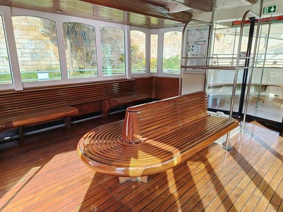 Intérieur du bateau en bois verni, avec un parquet, une banquette centrale double et arrondie en lattes de bois, des banquettes le long des vitres et une double porte vitrée qui mène à l'arrière du bateau. Le soleil donne une belle lumière et des ombres.