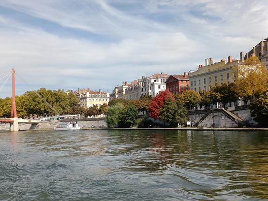 Vue prise de l'arrière du bateau, avec quelques vagues sur l'eau, un pont suspendu avec un hauban en métal rouge, des quais avec des arbres aux couleurs automnales et des bâtiments. Le ciel est lumineux mais voilé.