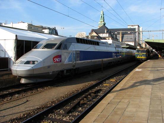 Une rame TGV gris argenté avec bandeau bleu en gare de Luxembourg-Ville. A l'arrière-plan, le bâtiment de la gare et à droite sur la voie 4, une locomotive SNCB en livrée bleu-vert avec bandes jaunes.

A TGV-trainset in silver grey livery with blue strip in Luxembourg-Ville, in the background the railway station buildings and right a SNCB/NMBS locomotive in blue-green with yellow strips.
