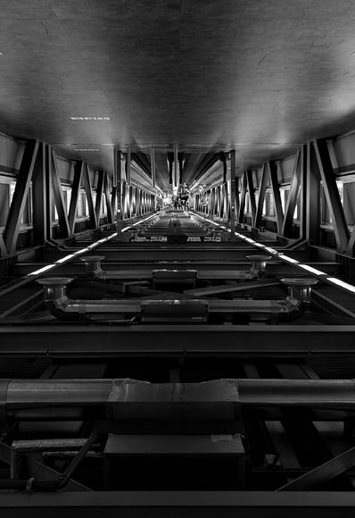 An upside down photo of a long corridor. The look is one of n industrial interior with metal beams, ducts, and pipes extends into the distance under a flat ceiling, illuminated by linear lights along the structure.