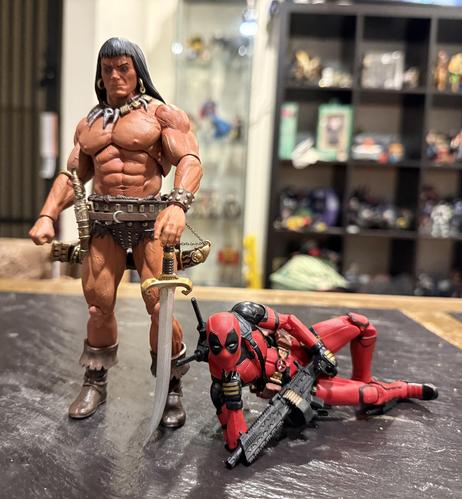 A 1:12 figure of Conan the Barbarian in the Frank Frazetta style leaning on his sword, glowering at the camera. A 1:12 Deadpool figure is stretched out at his feet in a sexy pose with a huge machine gun suggestively near his crotch.