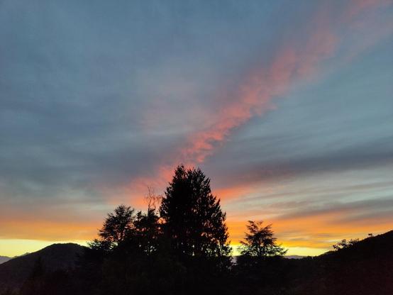 Photo du soleil couchant, dont les derniers rayons se reflètent sur un plafond nuageux. C'est très clair à l'horizon sur les montagnes du Vercors, puis rose, avec une lignée nuageuse qui traverse en diagonale, puis cela devient bleu-gris à mesure que l'on s'éloigne. Au premier plan, des collines et des arbres aux silhouettes déjà sombres.