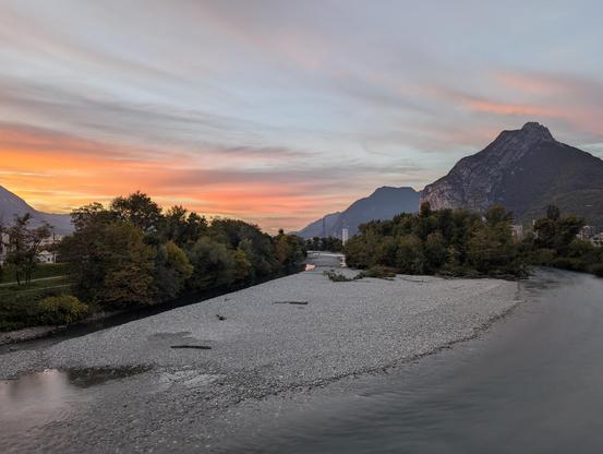Photo d'une rivière (le Drac) avec en arrière plan quelques arbres, des montagnes de part et d'autre, et le soleil couchant en arrière plan.
