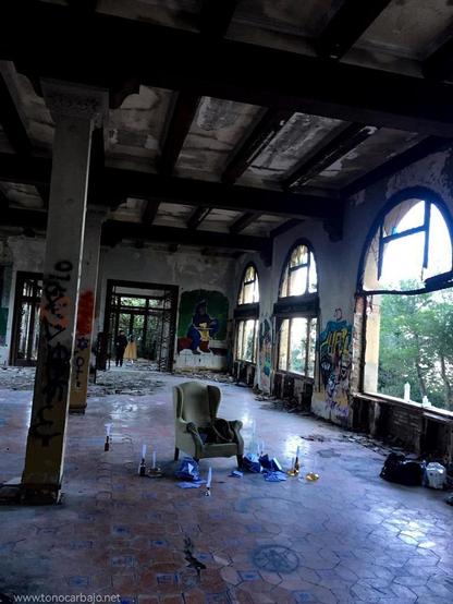 Una sala abandonada, con techos y paredes en ruinas, iluminada por grandes ventanales con arcos. En el centro, un sillón solitario rodeado de escombros, velas y grafitis. La luz natural entra en contraste con el deterioro, creando una atmósfera de melancolía y nostalgia.