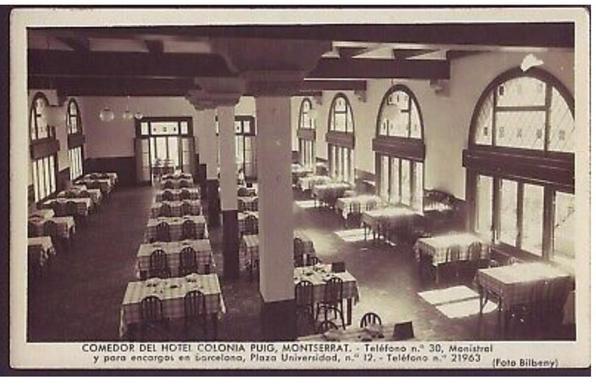 Una antigua postal del Comedor del Hotel Colonia Puig, en Montserrat, con mesas elegantemente preparadas y grandes ventanales que dejan entrar luz natural. Al pie, se indican datos de contacto: teléfono y dirección en Barcelona, Plaza Universidad, 12. La foto, en tonos sepia, evoca el ambiente clásico y sereno de mediados del siglo XX. (Foto Bilbeny)