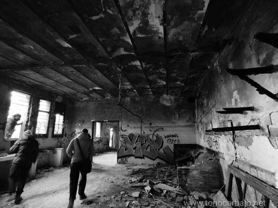 Un espacio abandonado, en blanco y negro, con el techo y las paredes deterioradas. Tres personas exploran entre escombros y grafitis, mientras la luz entra por ventanas rotas. La atmósfera transmite melancolía y el paso del tiempo