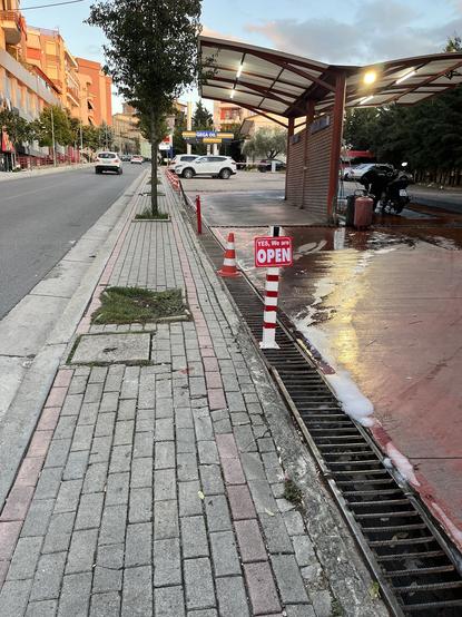 View from sidewalk made of pavers up a hill. An already-narrow sidewalk has small square street tree wells with young trees in them. The one nearest the car wash driveway is missing its tree, the stump underneath weeds that have filled the well. A man washes his car under the shelter of the self-car wash. Traffic cones over the grill between the private property and the sidewalk hold a sign saying in English "Yes, we are OPEN".