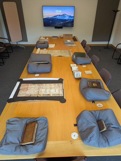 Leather bound volumes, manuscripts, positioned on grey cushions along a long wooden table. Several large scale facsimiles interspersed between original collection items.