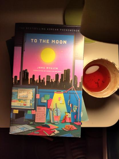 Kuvassa kirjan kansi, jossa värikäs, piirretty kuva työhuoneesta ja kaupunkimaisema sen ikkunasta. Taivaalla on täysikuu. Kirjan kannen lisäksi kuvassa on muki teetä sekä lamppu.