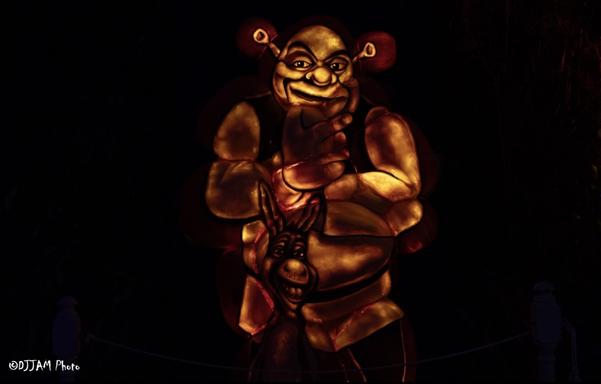 The image depicts a large, illuminated figure of a clown standing against a dark backdrop. The clown has a large, prominent head, and two small ears, giving it a distinctive appearance. The figure is holding a lantern in its hands and has an odd, human-like face.

In the foreground, there is a smaller, darker-colored figure, possibly a person or a part of the clown's outfit. A rope can be seen near the right edge of the image. The image has a text-based description, providing information about the size, color, and features of the clown figure.
