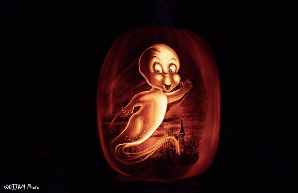 The image features a pumpkin carved to resemble a ghost. The ghost has a white body with black spots on its head and a round belly with a red outline. The pumpkin, glowing from within, casts a warm glow on the surrounding area, illuminating the scene in an eerie ambiance. The pumpkin is placed on a table, drawing the viewer's attention to its intricately carved design.