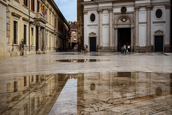 Español: Plaza con suelo de mármol mojado que refleja los edificios históricos a su alrededor. Al fondo se ve una iglesia blanca con columnas y un grupo de personas caminando.

Inglés: Square with wet marble floor reflecting the historic buildings around it. In the background, a white church with columns and a group of people walking.