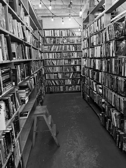 A cozy black and white bookstore aisle with shelves filled with books, a small stool, and string lights hanging from the ceiling.