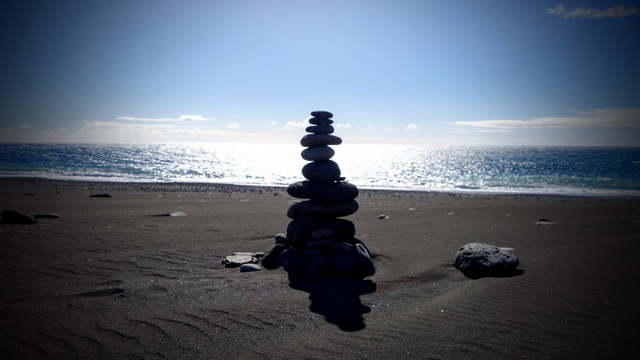 An einen Strand mit schwarzem Vulkansand hat jemand schwarze Kieselsteine aufeinander gestapelt. In Hintergrund glitzert der Atlantik in der tiefstehenden Nachmittagssonne silbern unter einem wolkenlosen Himmel. Der Steinturm wirft einen Schatten auf den Sand.