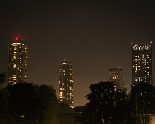 Four london tower blocks evenly spaced throughout the frame, with red lights on top, and the glow of the city below...there's also trees in the foreground