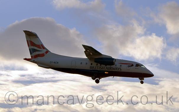 Side view of a high-winged, twin engined jet airliner flying from left to right at a low altitude, with undercarriage extended and flaps deployed from the rear of the wings, suggesting it is about to land.
The plane is largely white, with a blue belly, and black "British Airways" titles on the lower centre fuselage.
The engines slung under the wings are the same dark blue, and there is a twisted dark blue and red ribbon on the upper forward fuselage, behind the cockpit.
The tail is white, with a pair of wavy red lines with a wavy dark blue triangle between them.
Fluffy white clouds with patches of pale blue sky fill the rest of the frame.