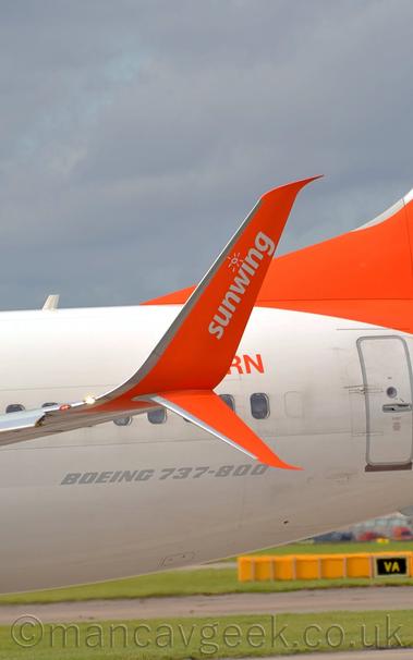 Close-up of the up- (and down-) turned wing tip of a twin engined jet airliner moving from right to left.
The plane is almost entirely white, with the last 2 letters "RN" of the registration on the upper rear fuselage, and grey text "Boeing 737-800" on the lower rear fuselage.
The wing-tips are bright orange, with white "Sunwing" titles under a stylised, child-like image of a sunburst.
The tail is the same bright orange.
Green grass lines both sides of a taxiway in the background, under the tail, with the rear of a yellow sign, and the front of a black sign with yellow letters "VA".
Grey sky with pale blue patches fill the rest of the frame.