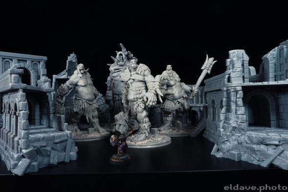 A photo of four unpainted 3d printed giants standing in the ruins of a multistory stone building, set against a black backdrop. There is a single knight figure in the foreground with their sword raised that barely reaches the knee of the giants. They're doing great.