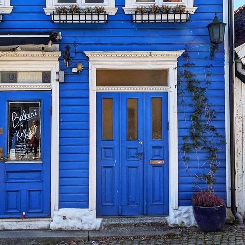 A blue-painted wooden building with two white-trimmed doors. The left door is for "Bakeri & Kafé." The main door on the right is a double door with four glass panels. A potted plant with a dead vine sits to the right of the door.