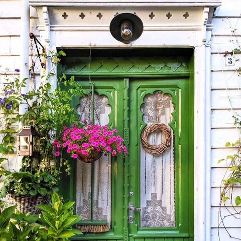 A bright green wooden double door with decorative window panes, surrounded by lush green plants and hanging baskets of pink petunias. A wicker wreath hangs on the left door.