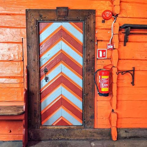A single wooden door painted with red and light blue chevrons, set in a rustic dark wood frame. The surrounding building is painted a vibrant orange. A fire extinguisher and fire alarm are visible on the right.