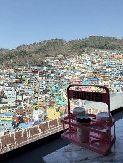 A vibrant cityscape with colorful houses, viewed from a cafe. A drink carrier with coffee cups sits on a table near the window.