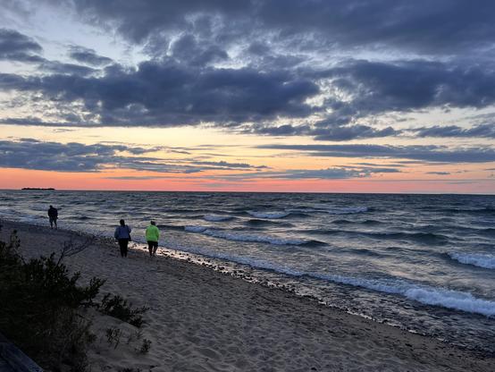 A serene beach scene at sunset featuring three people walking along the Lake Superior shoreline. The sky is filled with clouds and vibrant hues of orange, pink, and purple as the sun sets over the waves.