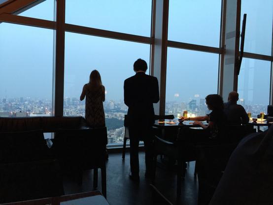 Silhouettes of people looking out the huge, tall windows on the top floors of the Park Hyatt Hotel in Tokyo. This level is a restaurant/bar, so some people are seated at tables, and others have gone to the window to look at the view of Tokyo. It is dusk, so the sky is blue and the lights of the city are just starting to turn on.