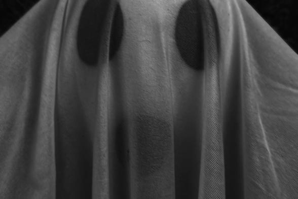 A black-and-white photograph shows a figure covered by a sheet, forming the outline of a human shape beneath. Three dark circular marks—two near the top, one below—suggest a ghostly face: eyes and a mouth, simple yet haunting. The lighting emphasizes the fabric’s folds and texture, creating a tactile rhythm of shadow and drape that makes the image feel eerie, minimal, and quietly surreal