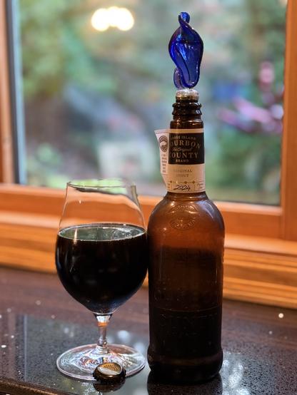 A snifter full of dark beer stands next to a tall brown bottle with a simple neck band label and a decorated cork stopper