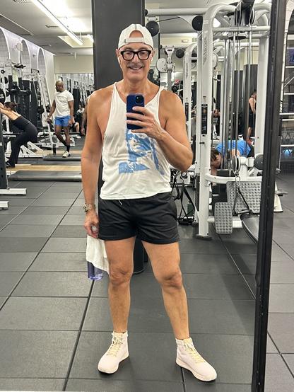 Me posing for a selfie in a gym, wearing a sleeveless white tank top, black shorts, and light pink sneakers. They are holding a water bottle and a towel. In the background, other gym-goers can be seen using workout.