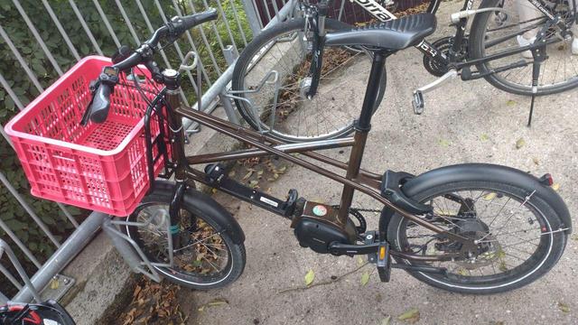 Cooper Bikes Uty E-Bike (mit herausgenommenem Akku) in Braun mit Frontgepäckträger und rosaroter Plastikkiste darauf steht in einem Fahrradständer.