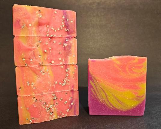 Atomic Sunrise soap. On the left are four bars stacked on their side to show the neon pink tops with some yellow and fuschia and star shaped biodegradable glitter. A single bar of soap sits on the right showing the feathered deaign