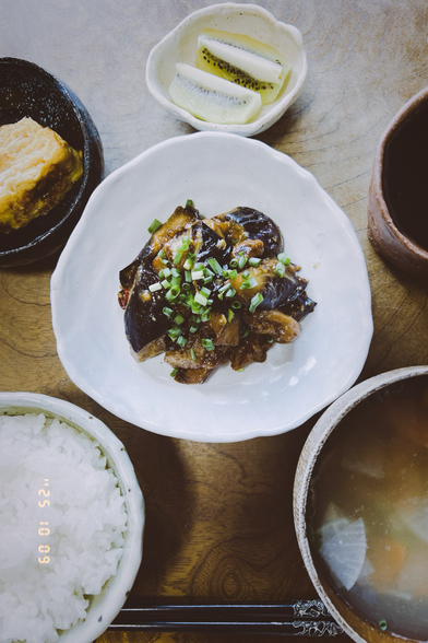 It’s stir-fried eggplant with miso sauce. It’s sprinkled with chopped green onion. It’s served on a triangle uneven white dish I made. Around the dish, there are some other dishes which have some foods.