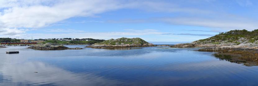 A panoramic photo of a body of water with small islands in it. The sky is blue with white clouds in it.