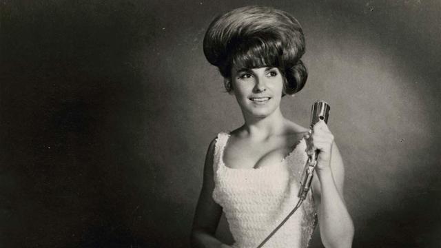 This is a sepia-coloured photo from the 1960s of Linda Lyndell.

It shows a young woman with an exaggerated bouffant hairstyle, in which the hair is combed back to be huge and swept across her head, culminating in a low bun on her left side.

Her eyes are turned to her left, she is smiling. 

She wears a white (or light-coloured) tight ruffled dress. It has a square neckline and accentuates her decolletage. Her arms are bare and she is holding, in her left hand, an old-fashioned microphone with a long cord attached.
