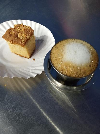 A square of cake and a foamy drink sit on a metallic surface.