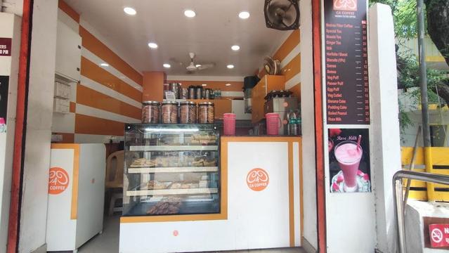 A small C4 Coffee shop with white and orange decor displays a menu board with prices. A glass cabinet holds pastries and jars.