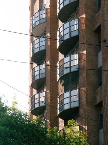 Image of a brick residential building featuring multiple rounded balconies. Green foliage is visible in the foreground, with power lines crossing the scene. The lighting indicates a bright, sunny day.