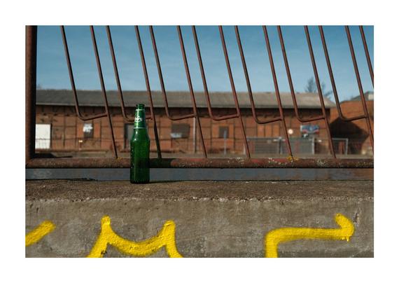 Eine grüne Bierflasche steht auf einer rauen Betonfläche im Vordergrund. Die Flasche ist leer und leicht von der Sonne beleuchtet, wodurch ihr grüner Farbton besonders hervorsticht. Hinter der Flasche verläuft ein rostiger Metallzaun mit vertikalen Streben, der das Bild in zwei Ebenen teilt. Dahinter befindet sich ein großes Industriegebäude mit Backsteinwänden und einem Dach aus gewelltem Blech. Die Architektur wirkt alt und verlassen, was dem Bild eine urbane, leicht melancholische Stimmung verleiht. Der Himmel ist klar und blau, was auf einen sonnigen Tag hindeutet. Auf der Betonfläche im unteren Bereich des Bildes ist gelbe Graffiti zu erkennen – teils verwischt, teils lesbar – was dem Ort eine zusätzliche Note von Verlassenheit und Street-Art-Charme verleiht. Die gesamte Szene wirkt wie ein Schnappschuss aus einem industriellen Randgebiet, das nicht mehr aktiv genutzt wird, aber dennoch Geschichten erzählt – von Arbeit, Alltag und urbaner Kultur.