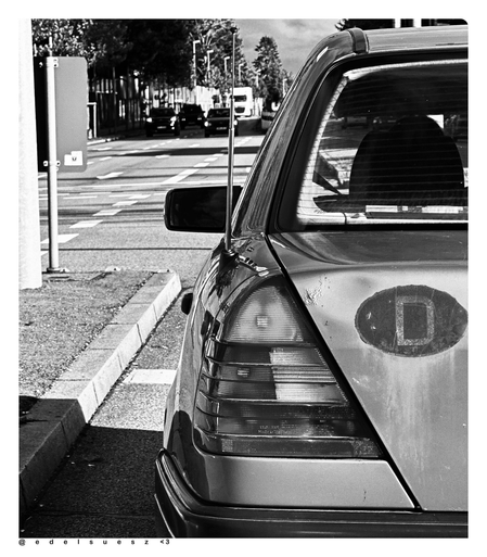 Schwarzweißfotografie: warten an einer Linksabbiegeampel, das Heck einer alten Mercedes Benz C Klasse voraus (1993–2001) im Mittelpunkt das linke, eckig zulaufende Rücklicht, daneben eine Delle im Kofferraumdeckel, darauf ein gealterter Kennzeichen D Aufkleber / Abziehbild, weit entfernter Gegenverkehr